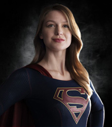 A woman with long blonde hair wearing a dark blue superhero suit with a red S emblem and a red cape.