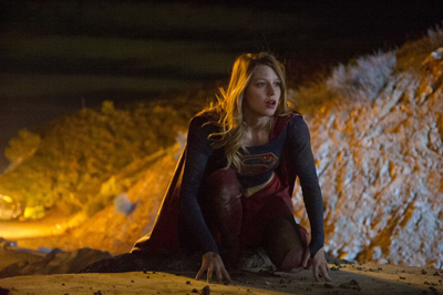 A blonde woman in a blue and red superhero suit crouches on a rocky slope under dim light.