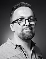 Black and white headshot of a man wearing thick-rimmed glasses and a button-down shirt.