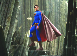 Superman in a blue suit and red cape stands atop jagged rock formations in a crystalline environment.