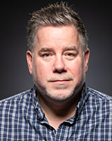 A man with short salt-and-pepper hair and a light beard wearing a blue and white plaid shirt.