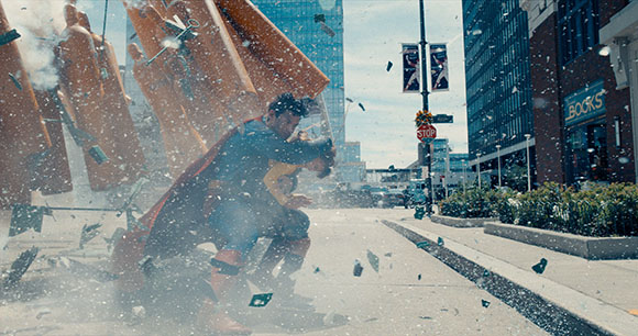 A superhero in a red cape crashes into a large orange structure on a city street amidst flying debris.