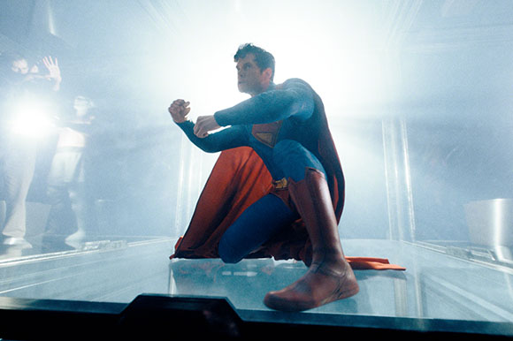 A man in a blue superhero suit and red cape crouches in a bright, misty room.