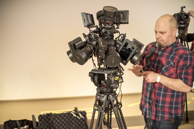A technician in a red plaid shirt adjusts a complex multi-camera rig mounted on a tripod.