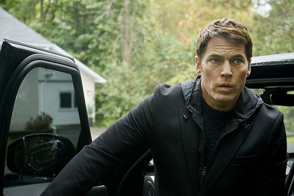 A man with short brown hair wearing a black jacket looks intensely to his left while standing by a car.