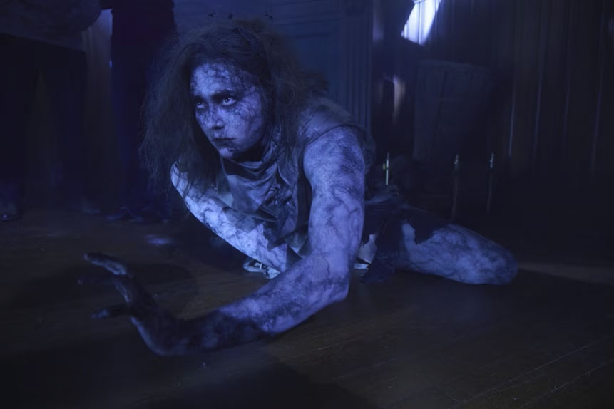 A person with dark, smudged skin crawls on all fours across a dimly lit wooden floor.