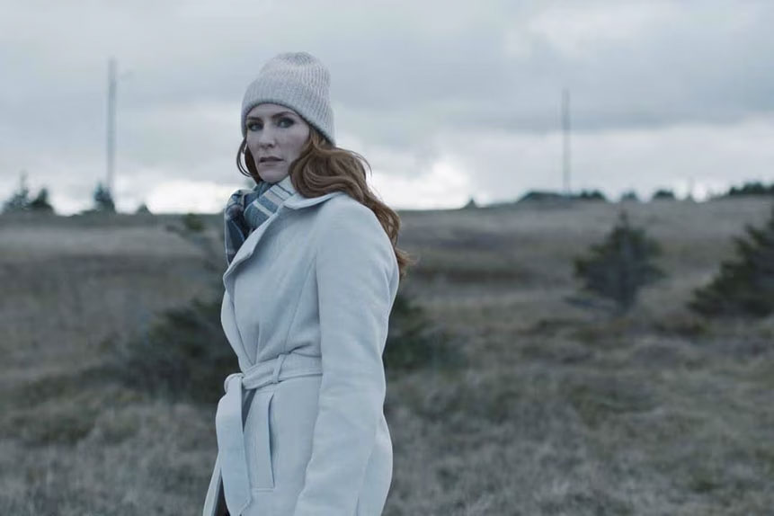 A woman with red hair wearing a white coat and light beanie stands in a grassy field under a cloudy sky.