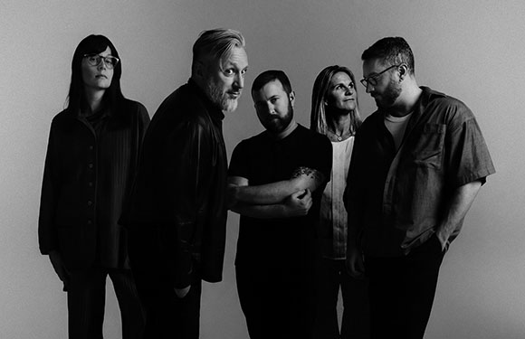 Five people stand in a group against a plain background in a black and white portrait.