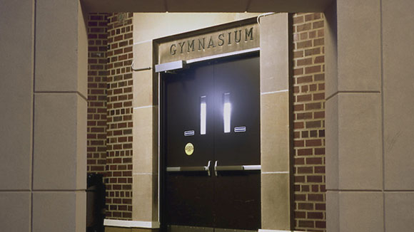 Dark double doors set in a brick and stone entryway with a sign reading GYMNASIUM above.
