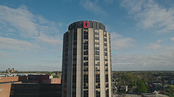 A tall, cylindrical skyscraper with a large red letter O on its roof against a cloudy blue sky.
