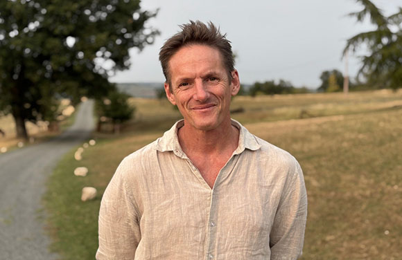 A smiling man with short brown hair wearing a light beige linen shirt in a field.