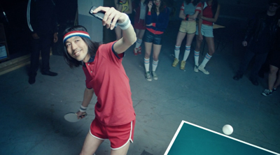 A man in a red shirt and headband holds up a phone to take a selfie next to a ping pong table.