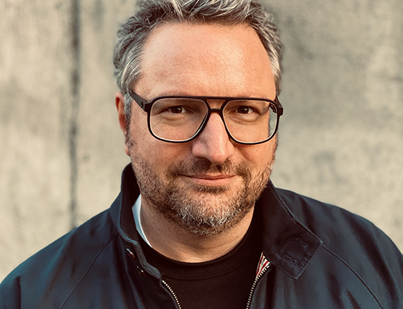 A man with salt-and-pepper hair and a beard wearing black-rimmed glasses and a dark jacket.