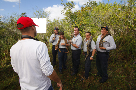 Six people stand in a field holding large snakes while a man in a red cap watches from the foreground.
