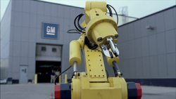 A yellow industrial robotic arm stands in the foreground with a GM building in the background.