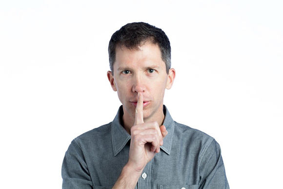 A man in a grey shirt holding his index finger up to his lips in a silence gesture against a white background.