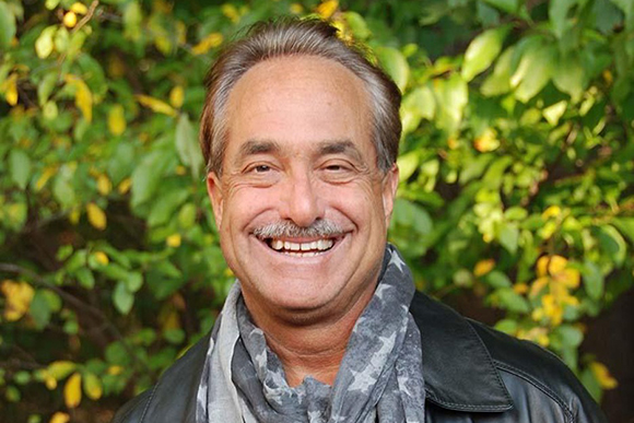 A smiling man with short brown hair wearing a black leather jacket and a patterned grey scarf.