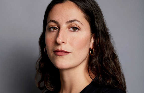 A woman with dark wavy hair and gold hoop earrings looks forward against a gray background.