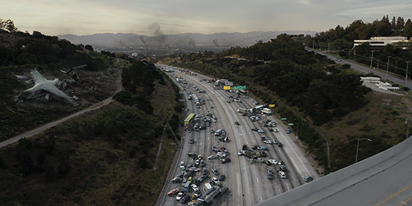 Aerial view of heavy traffic on a multi-lane highway winding through a hilly landscape under a cloudy sky.