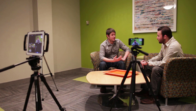 Two men sit in armchairs in a green room, talking to a camera on a tripod and a tablet on another tripod.