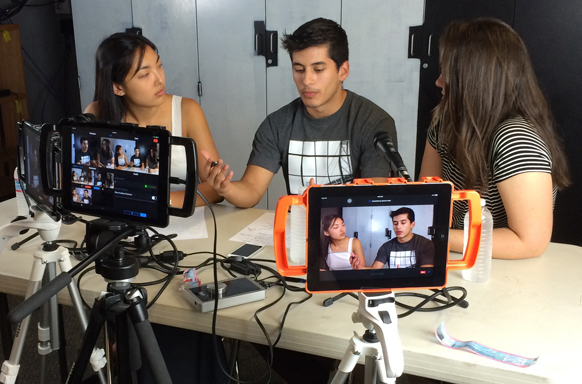 Three young adults sit at a table with cameras and a tablet displaying their video feed.