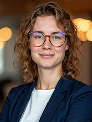 A smiling woman with curly blonde hair and round glasses wearing a navy blazer and white shirt.