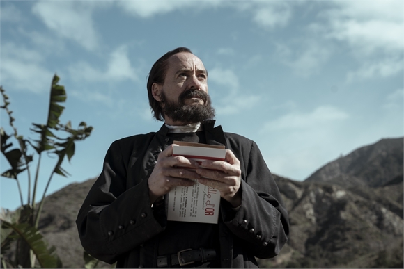 A bearded man in a black jacket holds a small rectangular box against a backdrop of mountains and sky.