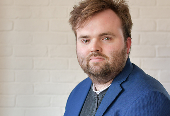 A man with brown hair and a beard wearing a blue blazer against a white brick wall.