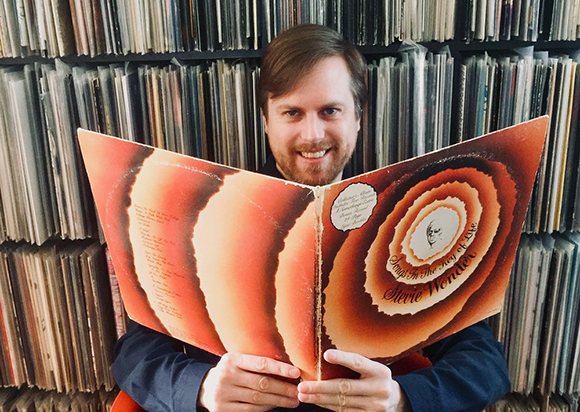 A man smiles while holding an open Stevie Wonder album cover in front of shelves of vinyl records.