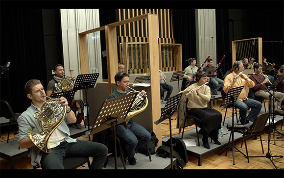 Musicians playing French horns, flutes, and clarinets during a rehearsal in a studio.