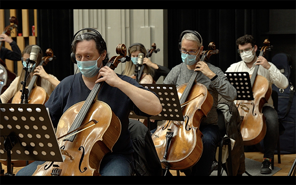 Musicians in face masks playing cellos in an orchestra setting.