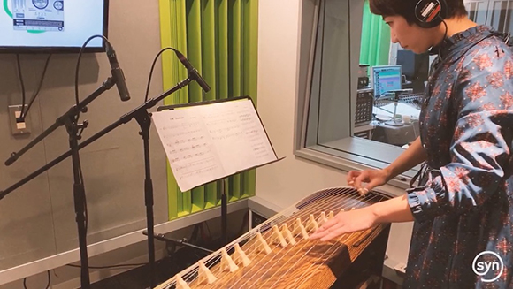 A person wearing headphones plays a long, wooden stringed instrument while looking at sheet music.