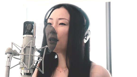 A woman with long dark hair wearing headphones sings into a professional microphone with a pop filter.