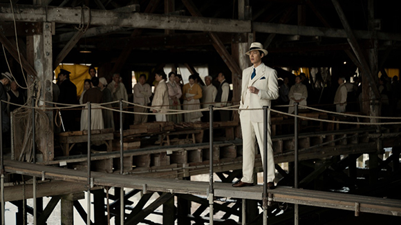 A man in a white suit and straw hat walks on a wooden pier with a crowd of people in the background.