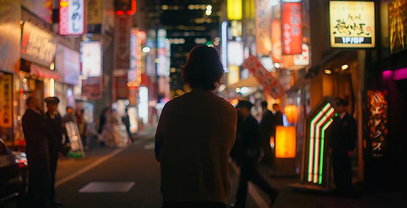 A person seen from behind walking through a narrow city street lined with glowing neon signs at night.