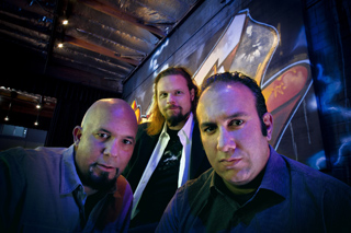 Three men looking at the camera in a dimly lit room with a large graffiti mural in the background.