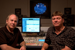 Two men sit in a recording studio behind a large mixing console and computer monitor.