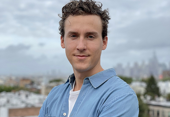 A young man with curly brown hair wearing a light blue button-down shirt against a blurred cityscape.