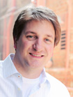 A smiling man with brown hair wearing a light blue collared shirt.