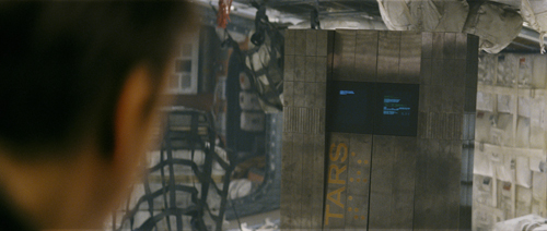 A person in the foreground looks toward a large, metallic rectangular robot with the word TARS on its side.