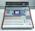 A silver audio mixing console with numerous faders, buttons, and a small central digital screen.