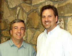 Two men smiling while standing side by side in front of a textured stone wall.