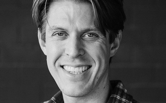 A black and white close-up portrait of a man with medium-length hair smiling at the camera.