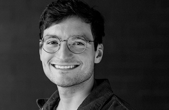 Black and white headshot of a smiling man wearing round-rimmed glasses.