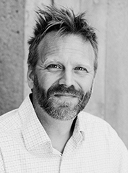 Black and white headshot of a man with short, messy hair and a beard, wearing a light-colored button-down shirt.