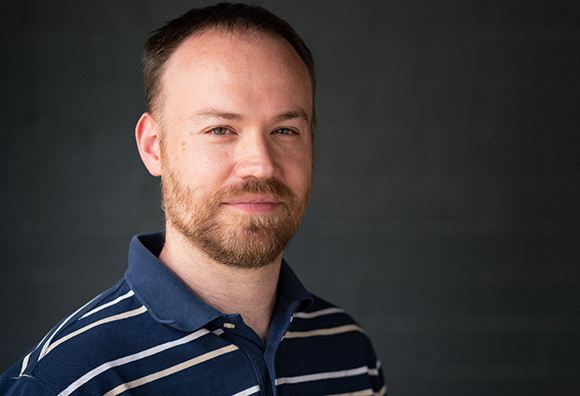 A man with short brown hair and a beard wearing a navy blue and white striped polo shirt.