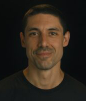 A man with short dark hair and a slight smile looking toward the camera against a black background.