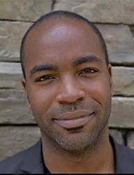 A smiling Black man with short hair and light facial stubble looking toward the camera.