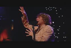 A person with light hair singing into a microphone with one arm raised against a dark background with lights.