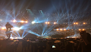 Bright spotlights beam across a massive, crowded stadium at night.